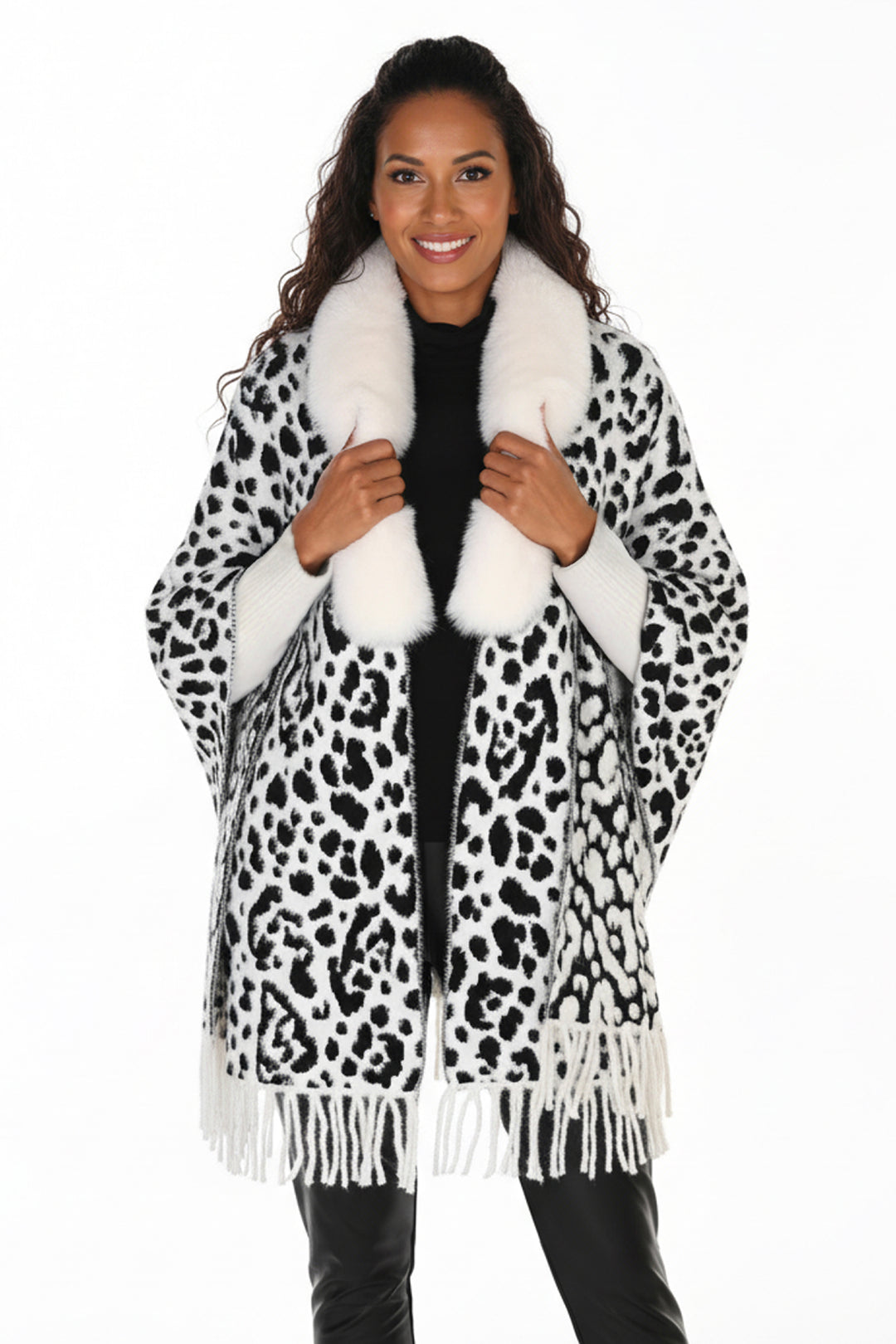 A woman modeling a black and white leopard print wrap coat with a fluffy white faux fur collar and fringe trim, worn over a black top and black pants against a plain white background.