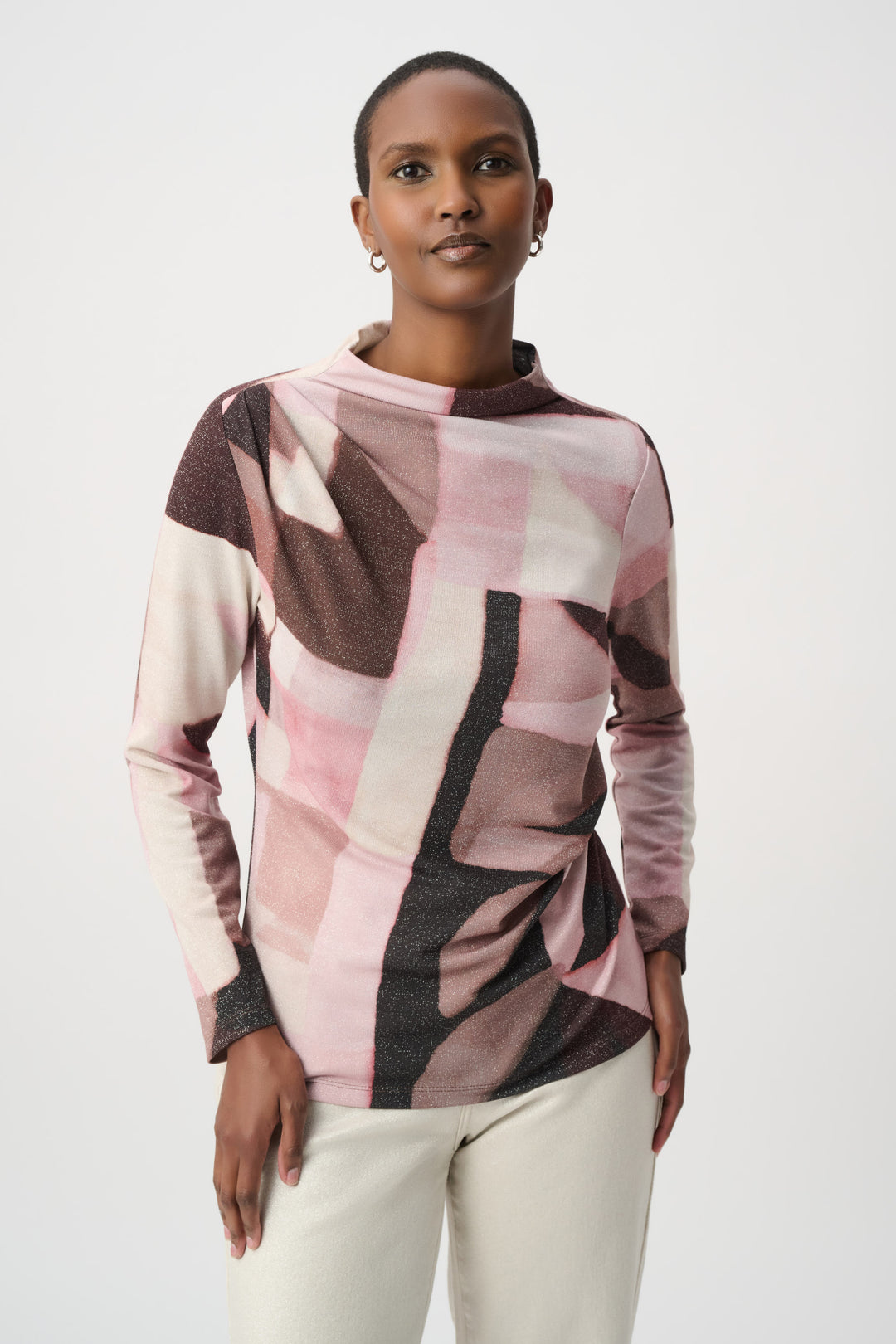 A woman wearing a long-sleeve mock-neck top with a pink, beige, and dark brown geometric block pattern, paired with cream pants, against a light gray studio background.