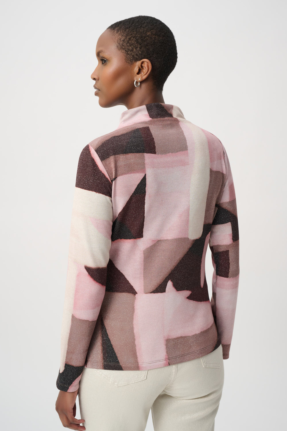 A woman shown from the back wearing a long-sleeve mock-neck top with a pink, beige, and dark brown geometric block pattern, paired with cream pants, against a light gray studio background.