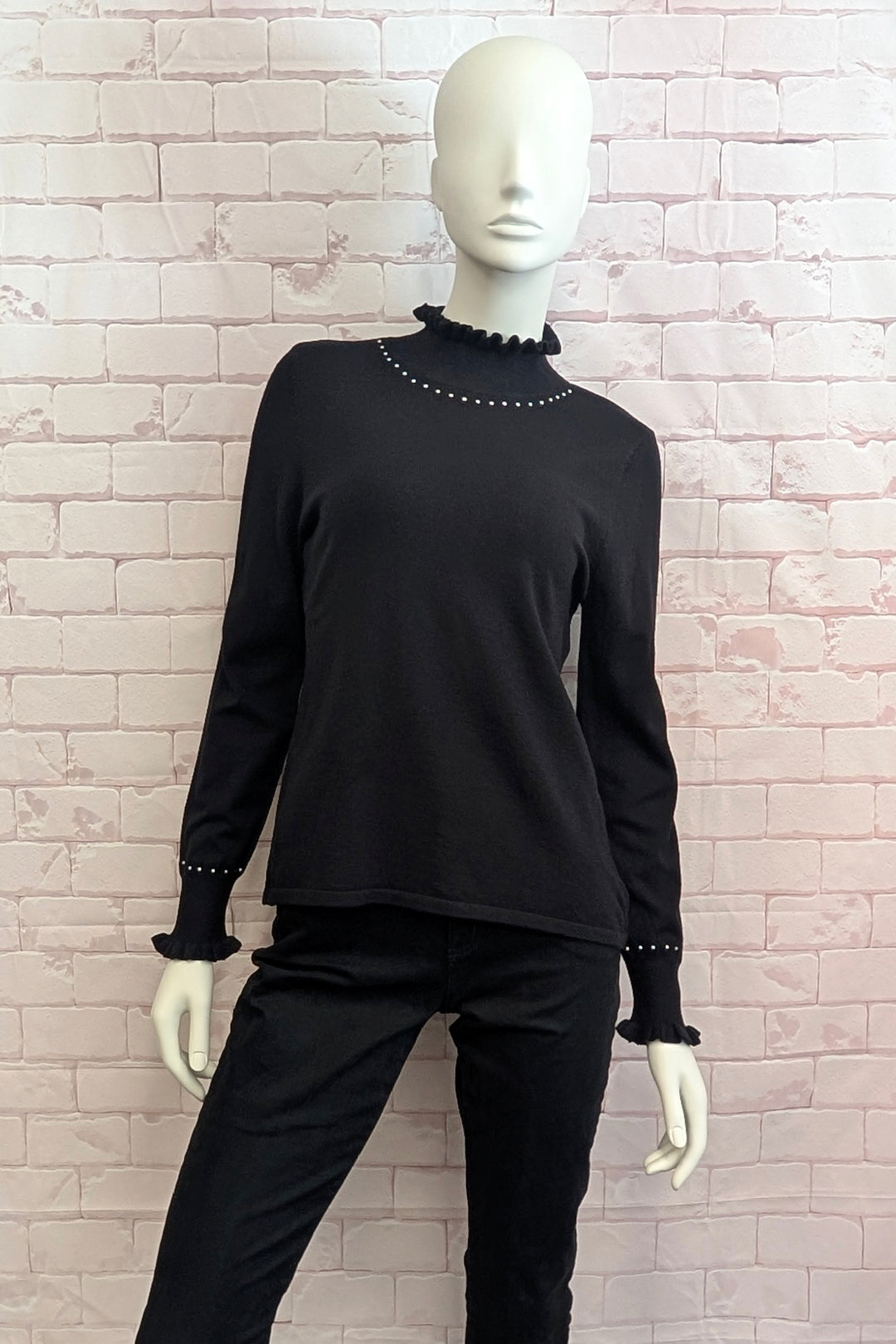 A black long-sleeve knit top with a ruffled mock neck and matching ruffled cuffs, displayed on a white mannequin against a light pink brick wall backdrop.