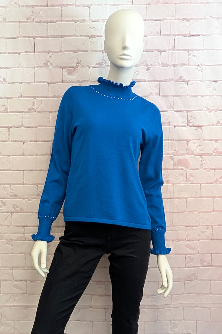 A bright blue long-sleeve knit top with a ruffled mock neck and matching ruffled cuffs, displayed on a white mannequin against a light pink brick wall. The top is styled with black pants.