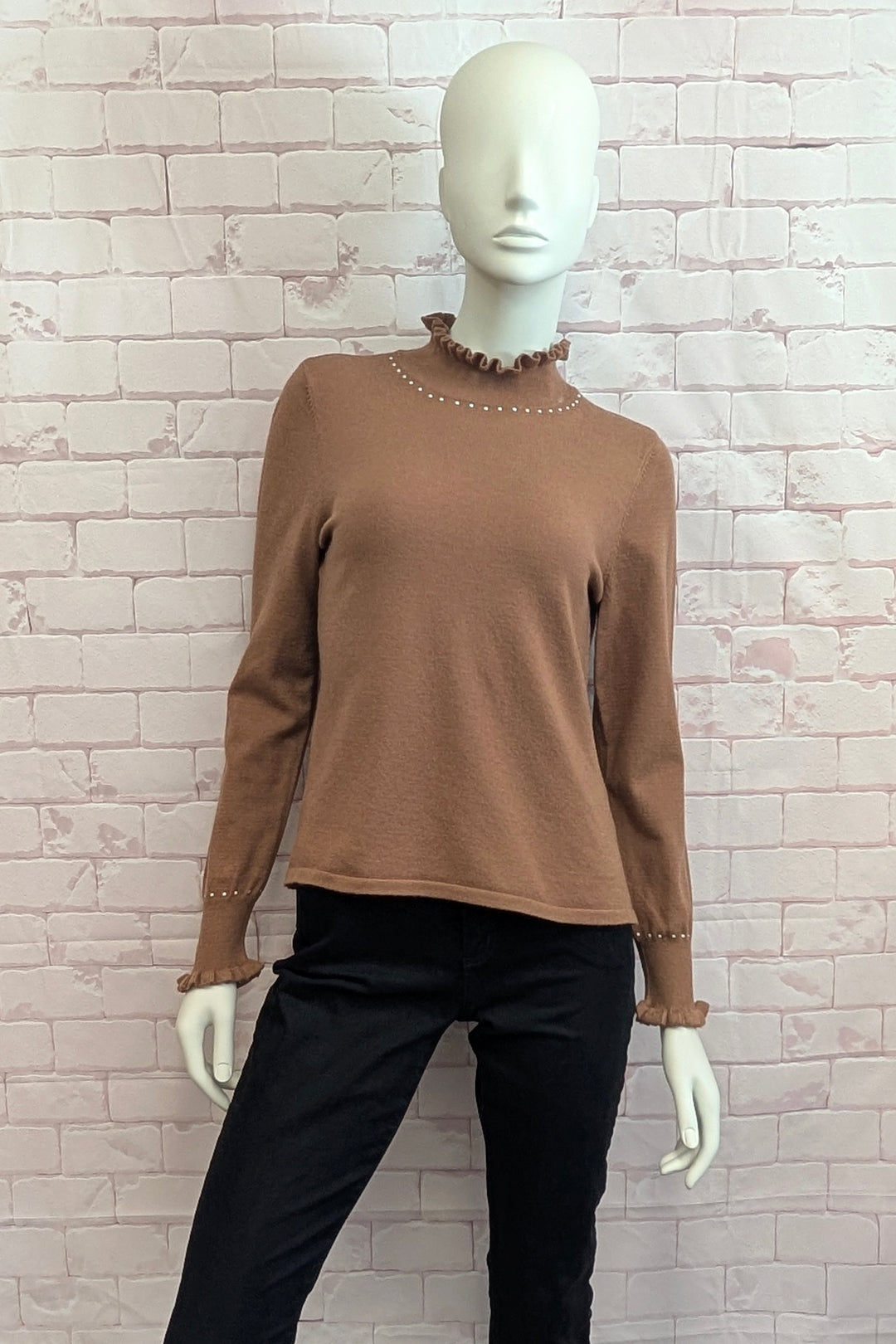 A brown long-sleeve knit top with a ruffled mock neck and matching ruffled cuffs, displayed on a white mannequin against a light pink brick wall. The top is styled with black pants.