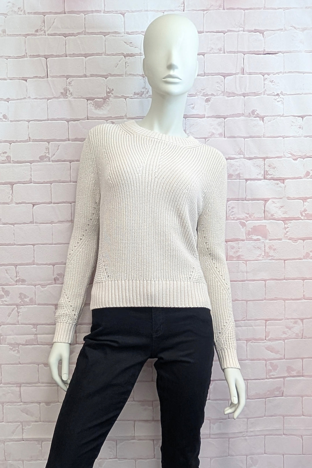 A mannequin wearing a ecru ribbed knit sweater with long sleeves and a crew neck, displayed on pink brick background.