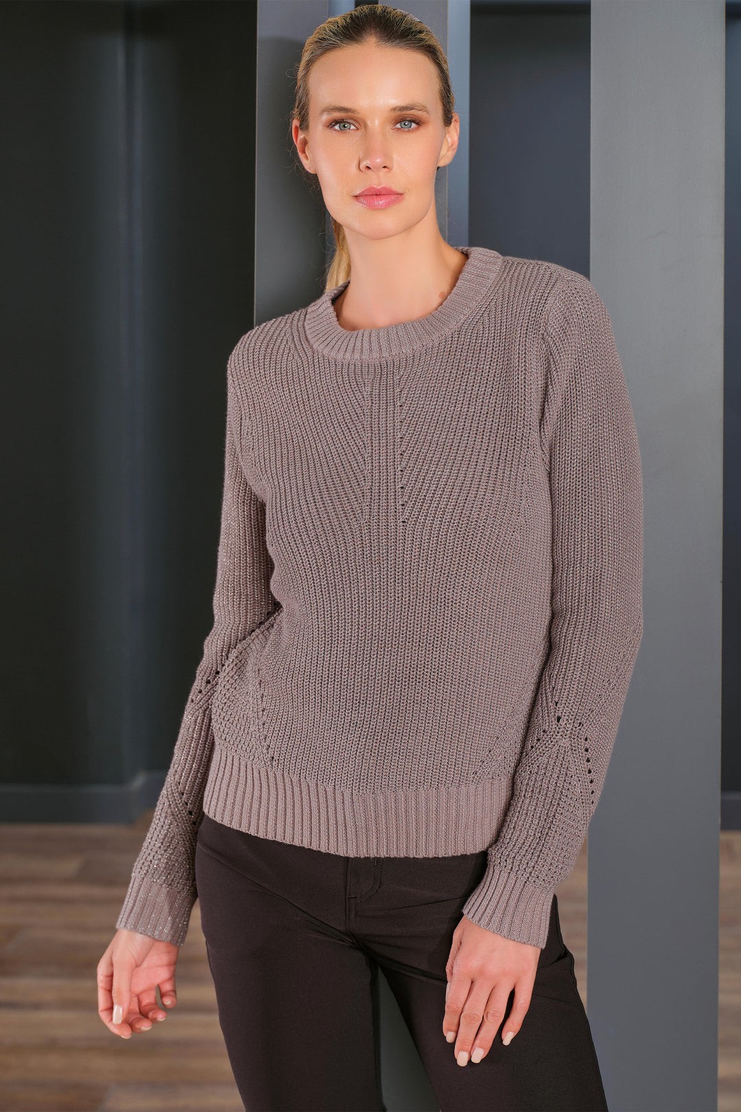 A woman modeling a taupe ribbed knit crewneck sweater with decorative openwork details on the sleeves, paired with black pants, set against a dark gray modern interior background.