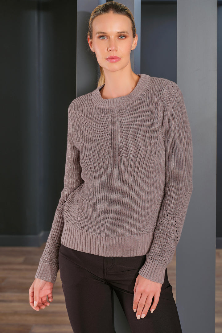 A woman modeling a taupe ribbed knit crewneck sweater with decorative openwork details on the sleeves, paired with black pants, set against a dark gray modern interior background.