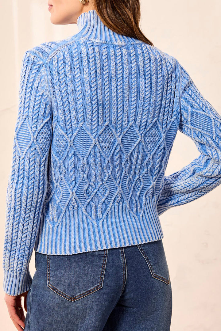 A woman wearing a light blue and white cable-knit mock neck sweater, shown from the back, paired with blue jeans against a neutral beige background.