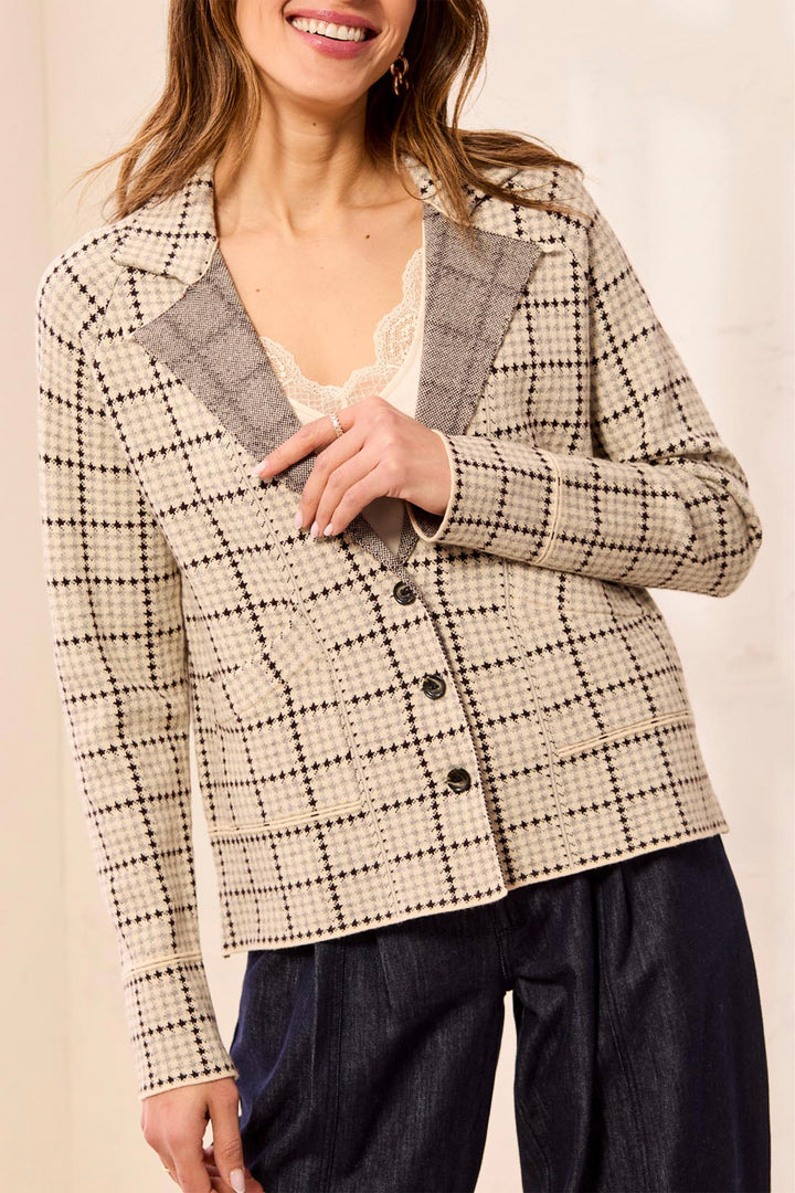 A woman wearing a beige and black plaid blazer with a gray textured lapel, paired with dark blue jeans and a cream lace top, shown against a warm beige background.