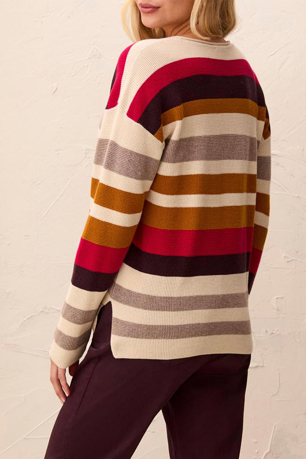 A woman wearing a striped knit sweater and dark plum pants, photographed from the back against a textured off-white wall.