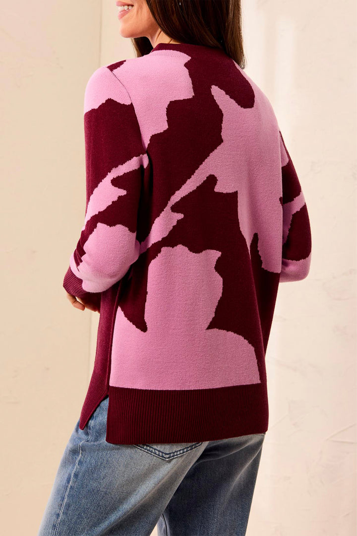 A woman wearing a pink and burgundy knit sweater with an abstract pattern, paired with light blue jeans, shown from a side-back angle against a neutral beige background.