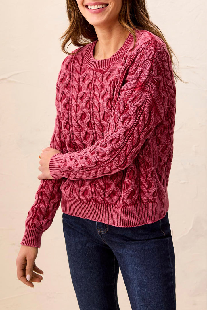 A woman wearing a chunky cable-knit sweater in a dusty rose pink color, paired with dark blue jeans, against a light beige textured wall.