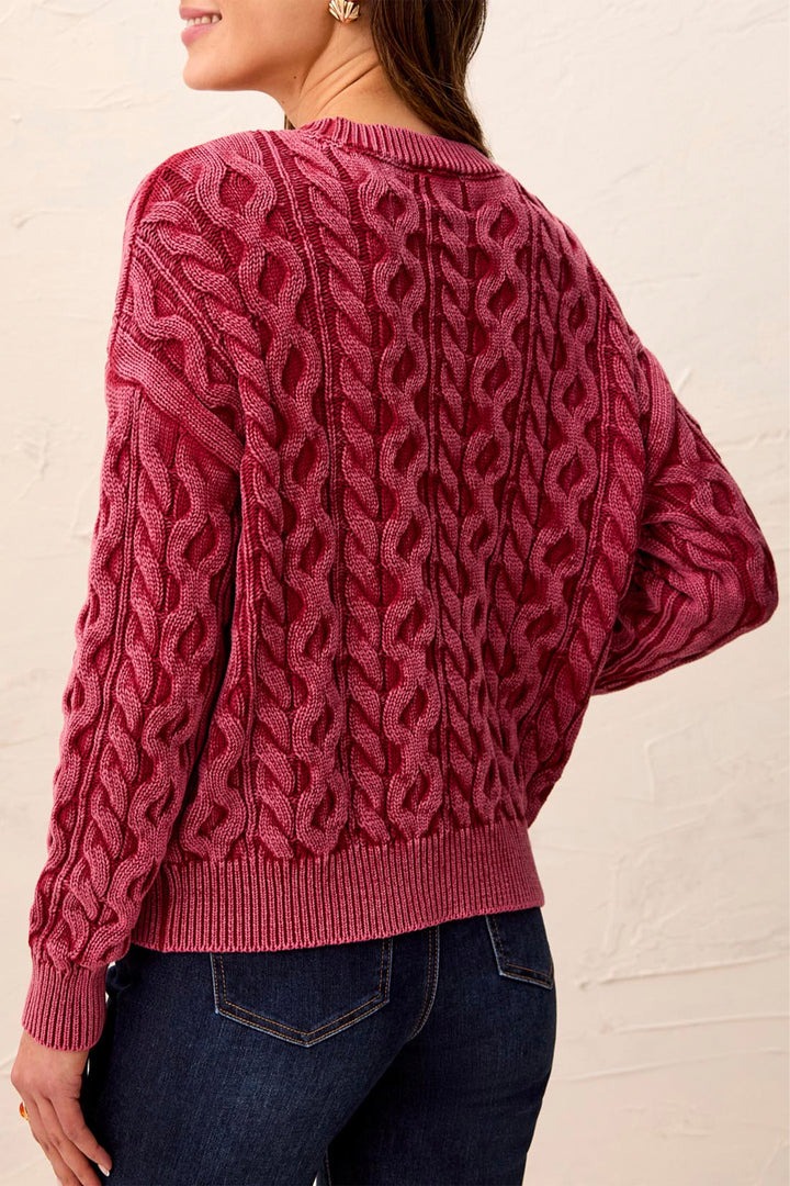 A woman wearing a chunky cable-knit sweater in a dusty rose pink color, shown from the back against a textured off-white wall.