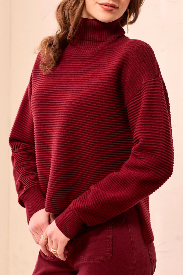 A woman wearing a ribbed, long-sleeve turtleneck sweater in deep burgundy, paired with matching pants, against a soft beige background.