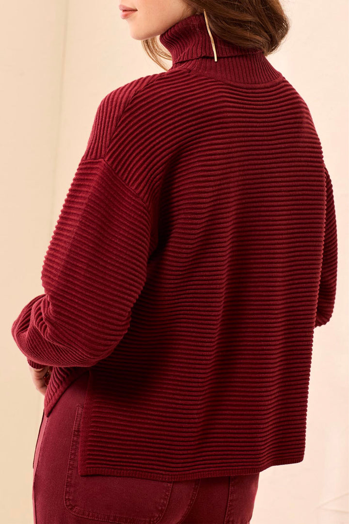 A woman wearing a ribbed knit turtleneck sweater in deep burgundy, shown from a side-back angle against a plain light beige background.