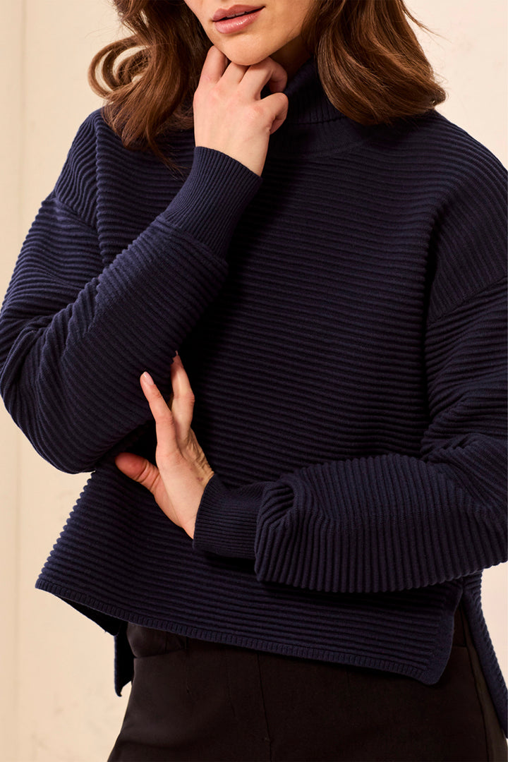 A woman wearing a ribbed knit turtleneck sweater in deep navy against a plain light beige background.
