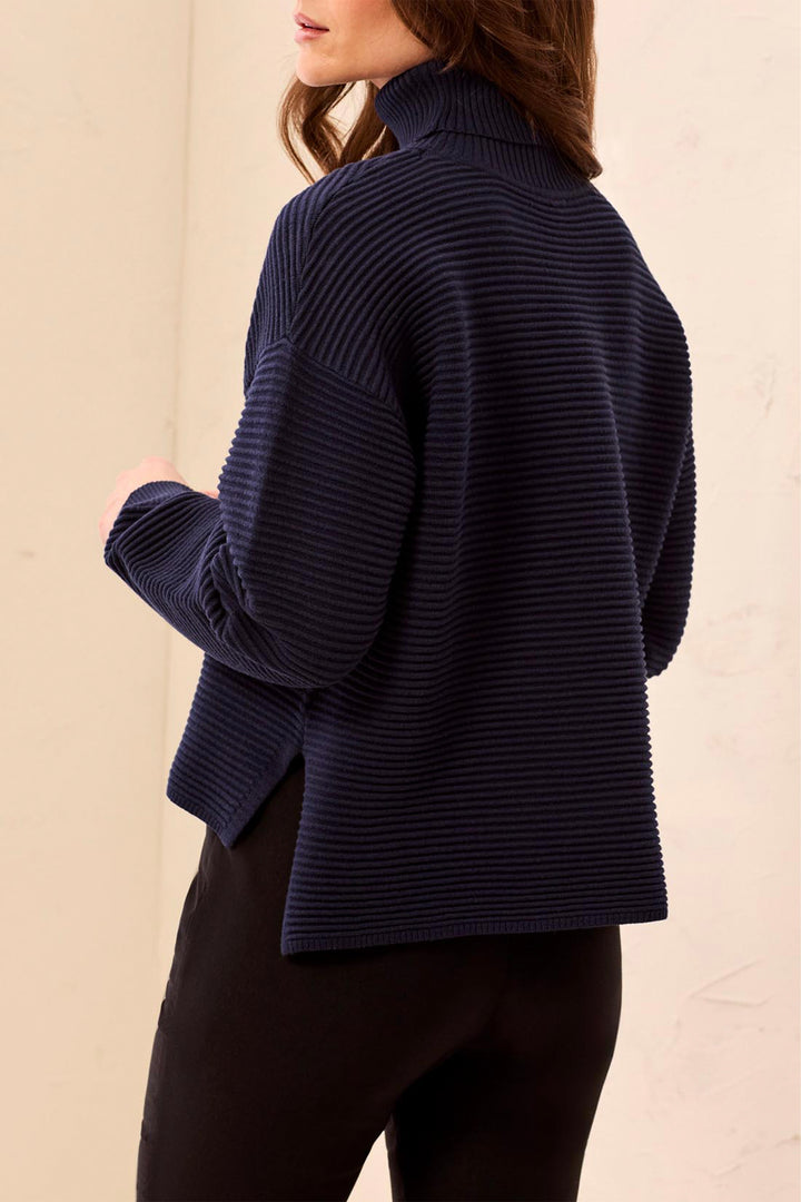 A woman wearing a ribbed knit turtleneck sweater in deep navy, shown from a side-back angle against a plain light beige background.