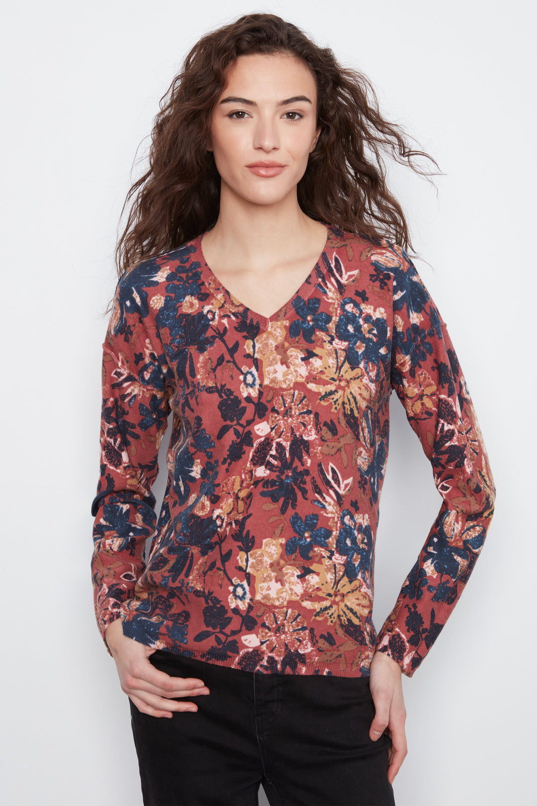A woman modeling a long-sleeve V-neck blouse with a dense floral print in rust red, navy blue, beige, and muted orange, paired with black pants against a light gray studio background.