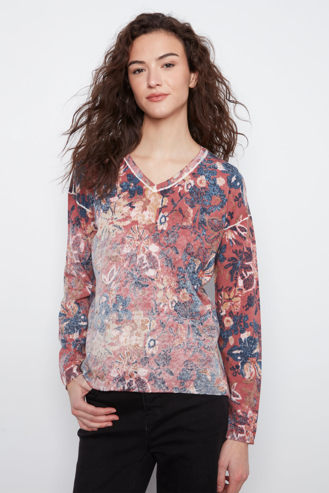 A woman with long, wavy brown hair wearing a long-sleeve V-neck top with a multicolor floral and botanical pattern in dusty pink, blue, and cream, paired with black pants, against a plain white studio background.