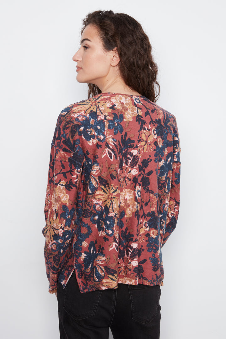 A woman shown from the back wearing a long-sleeve blouse with a dense floral print in rust red, navy blue, beige, and white, paired with black pants, against a plain white background.