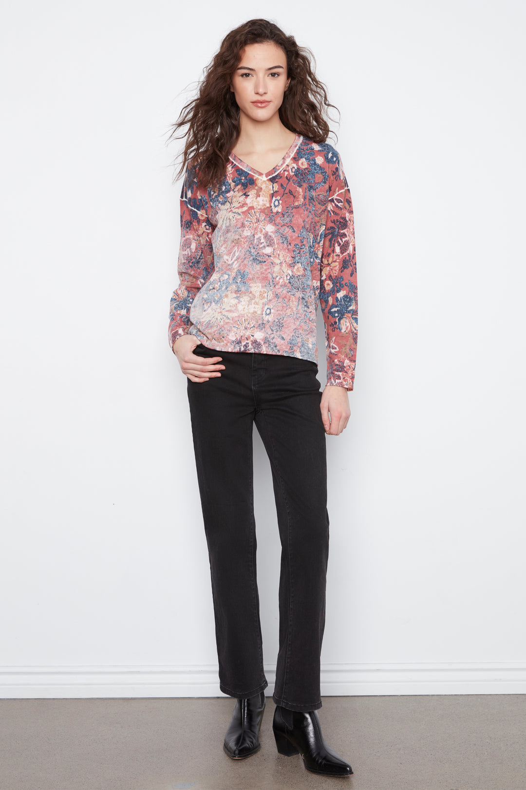 A woman stands against a plain white wall wearing a long-sleeve V-neck blouse with a dense floral and botanical print in rust red, navy, and beige tones, paired with black pants and black ankle boots.