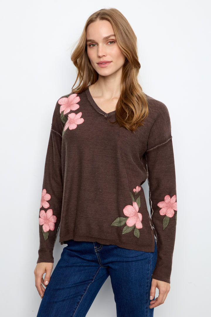 A woman wearing a brown long-sleeve V-neck sweater with pink floral embroidery on the shoulders and sleeves, paired with blue jeans, against a plain white background.