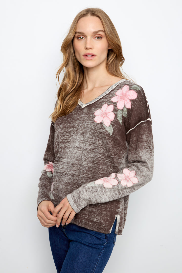 A woman wearing a brown and gray marled knit sweater with pink floral embroidery on the shoulders and sleeves, paired with blue jeans, against a plain white background.
