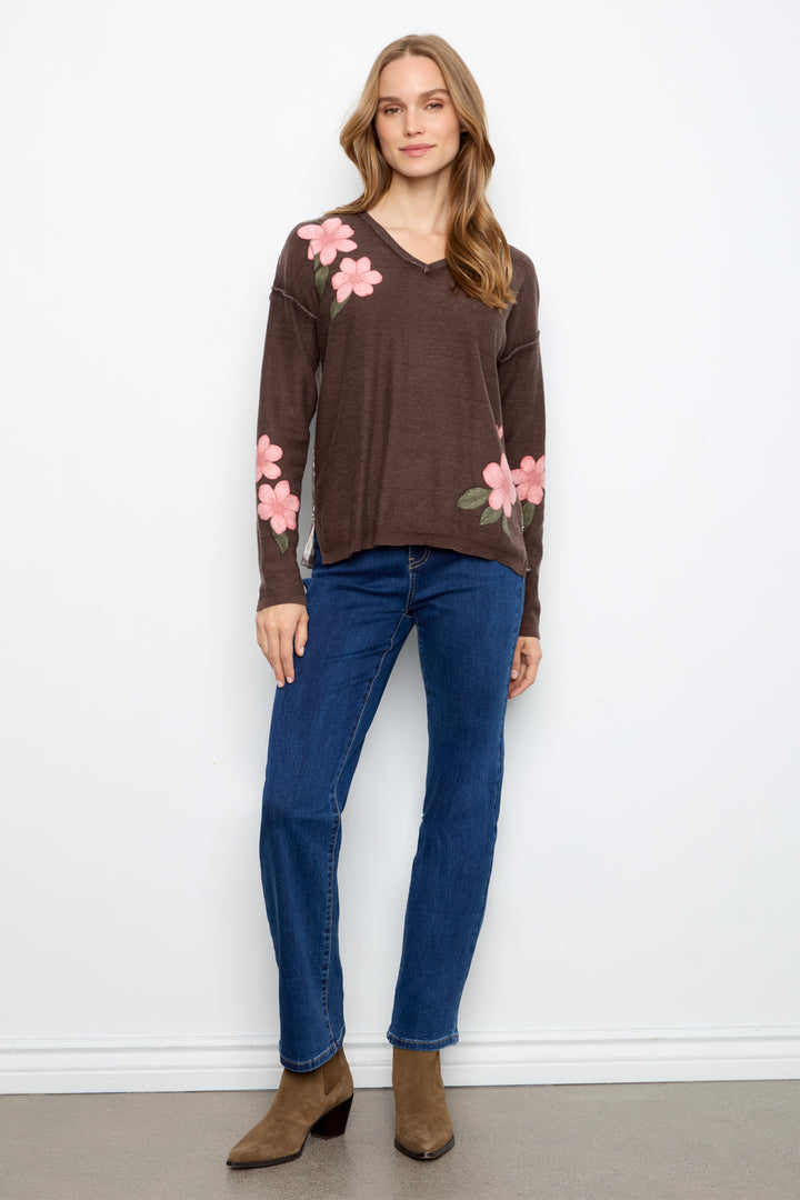 A woman modeling a brown long-sleeve V-neck sweater with pink floral embroidery on the shoulders and sleeves, paired with blue jeans and brown ankle boots, standing against a white wall.