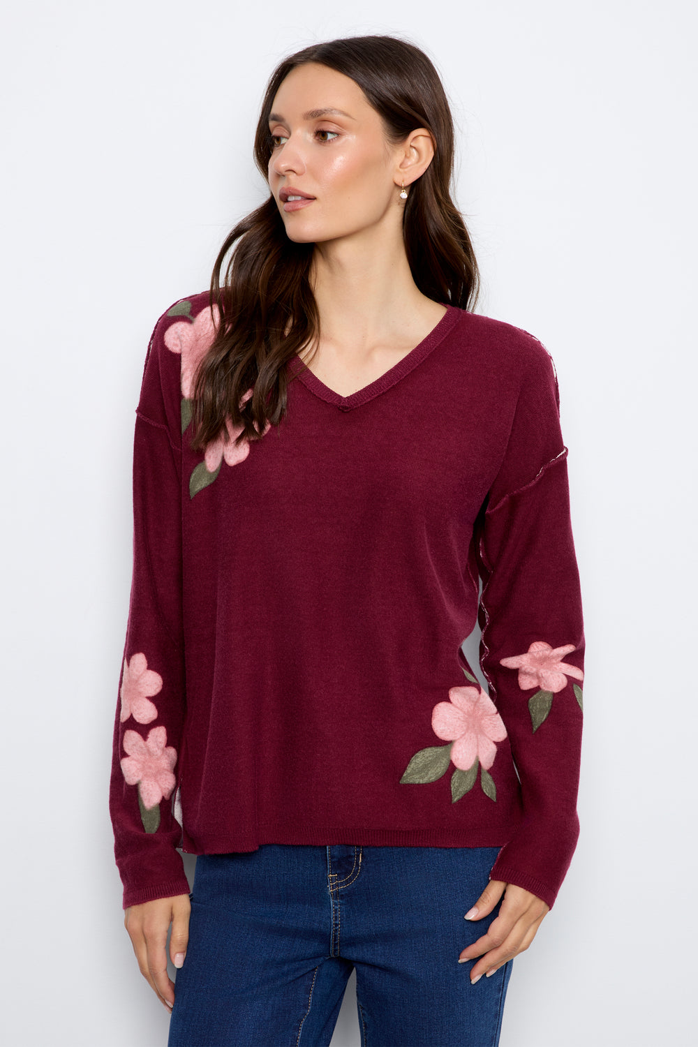 A woman wearing a maroon long-sleeve V-neck sweater with pink floral embroidery on the shoulders and sleeves, paired with blue jeans, against a plain light gray background.