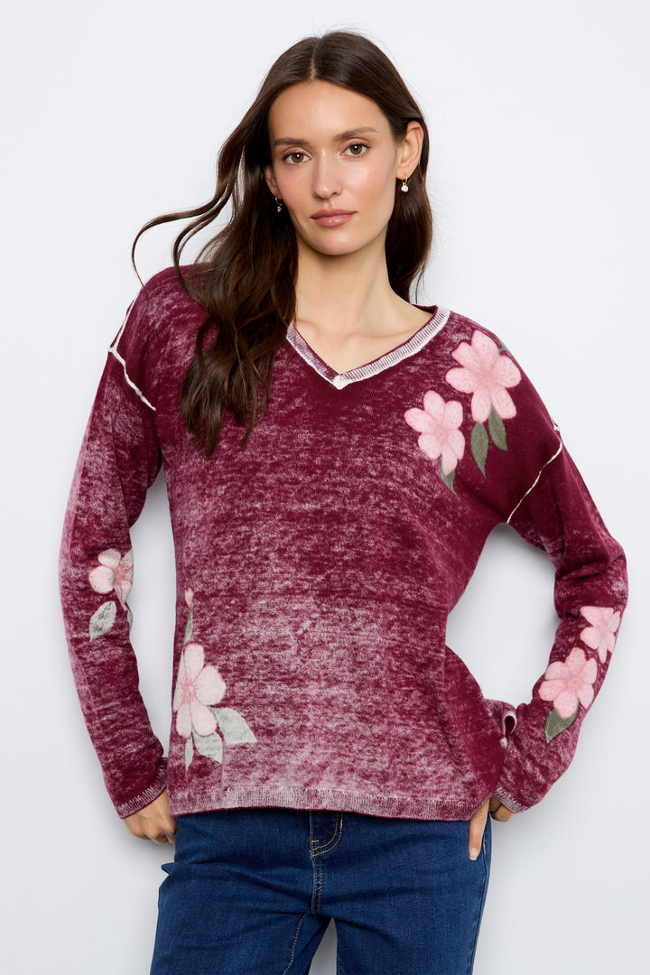 A woman modeling a maroon long-sleeve V-neck sweater with pink floral embroidery and light piping details, paired with blue jeans against a light gray background.