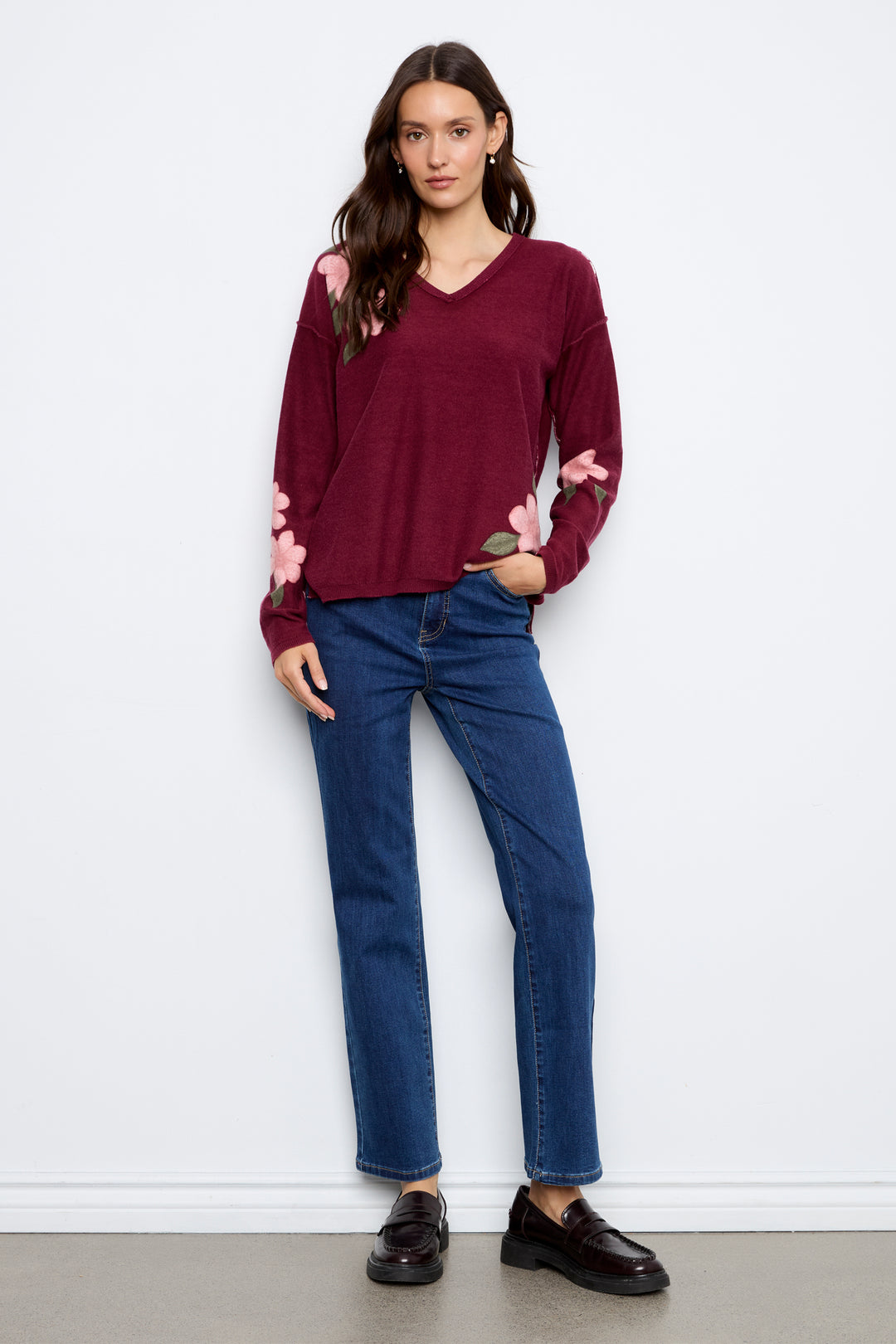 A woman modeling a burgundy long-sleeve knit top with pink floral embroidery on the shoulders and sleeves, paired with dark blue jeans and brown leather loafers, standing against a white wall.
