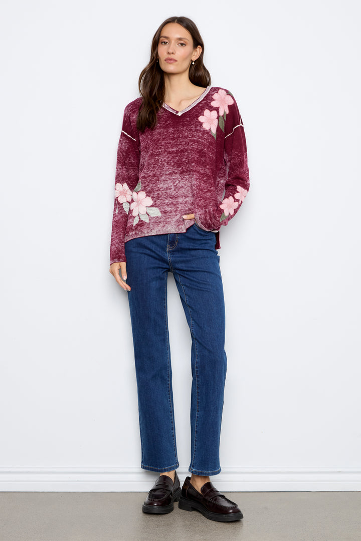 A woman stands against a white wall wearing a burgundy knit sweater with pink floral embroidery, blue jeans, and dark brown loafers.