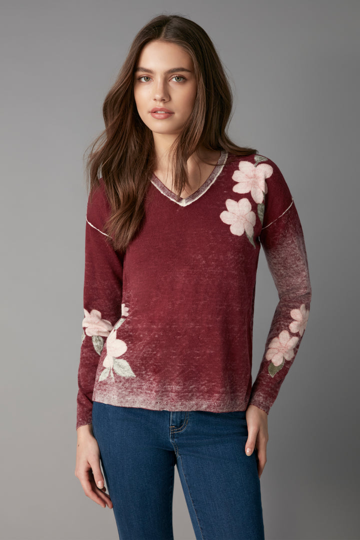 A woman modeling a long-sleeve V-neck sweater in a washed burgundy color with pink floral applique on the chest and sleeves, paired with blue jeans against a solid gray studio background.