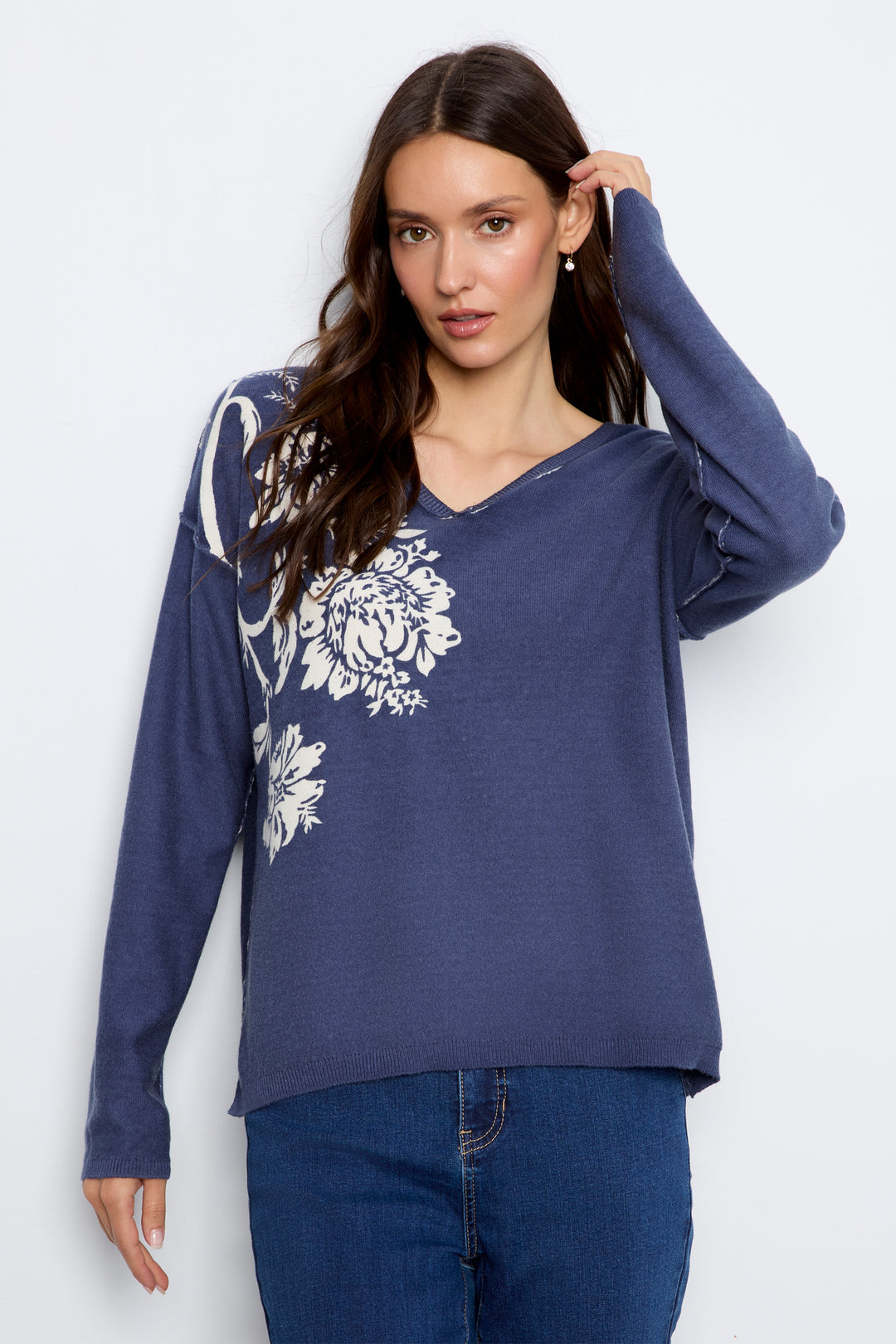 A woman modeling a navy blue long-sleeve V-neck top with a white floral print on the front, paired with blue jeans, against a light gray studio background.