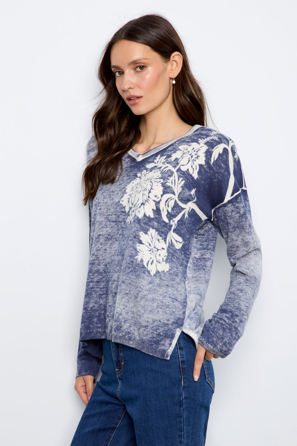 A woman modeling a long-sleeve V-neck top with a blue and gray marbled wash and a white floral print on the front, paired with blue jeans against a light gray studio background.