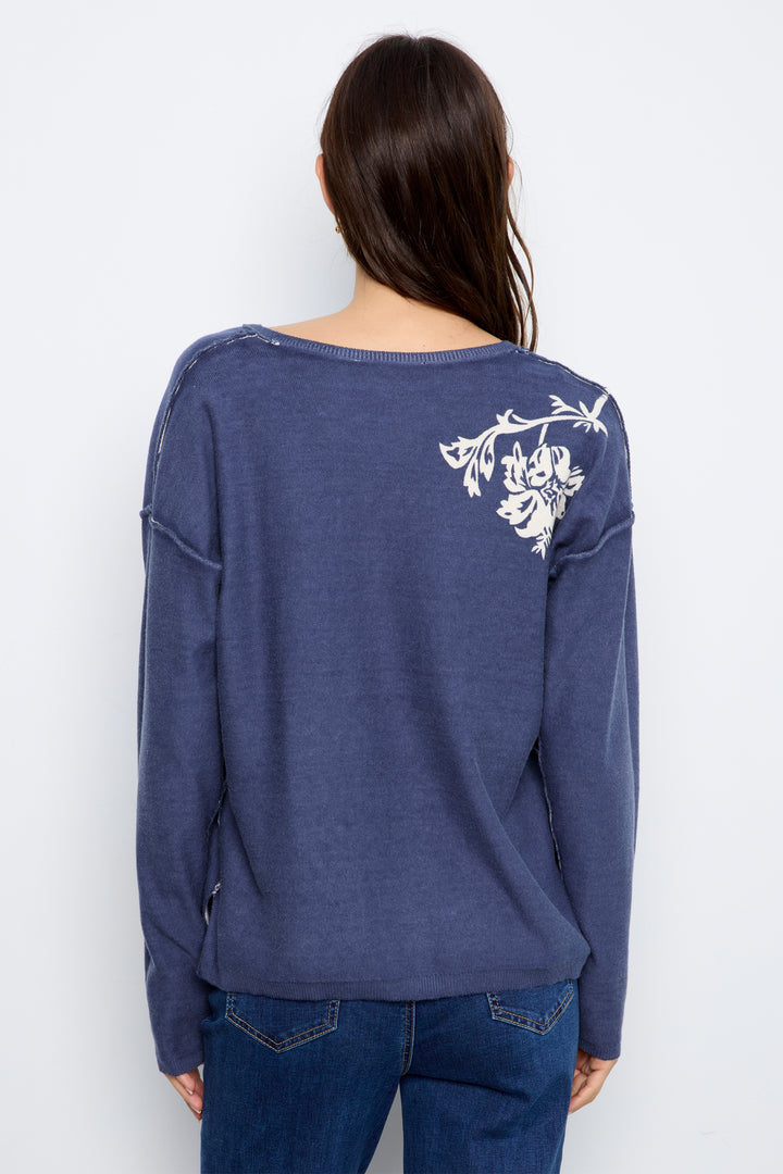A woman viewed from the back wearing a slate blue long-sleeve top with a white floral embroidery on the upper right shoulder, paired with blue jeans, against a plain white background.