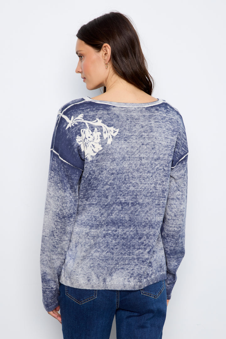 A woman shown from the back wearing a long-sleeve knit top with a blue and white mottled gradient and a white floral graphic on the upper left shoulder, paired with blue jeans against a plain light gray background.