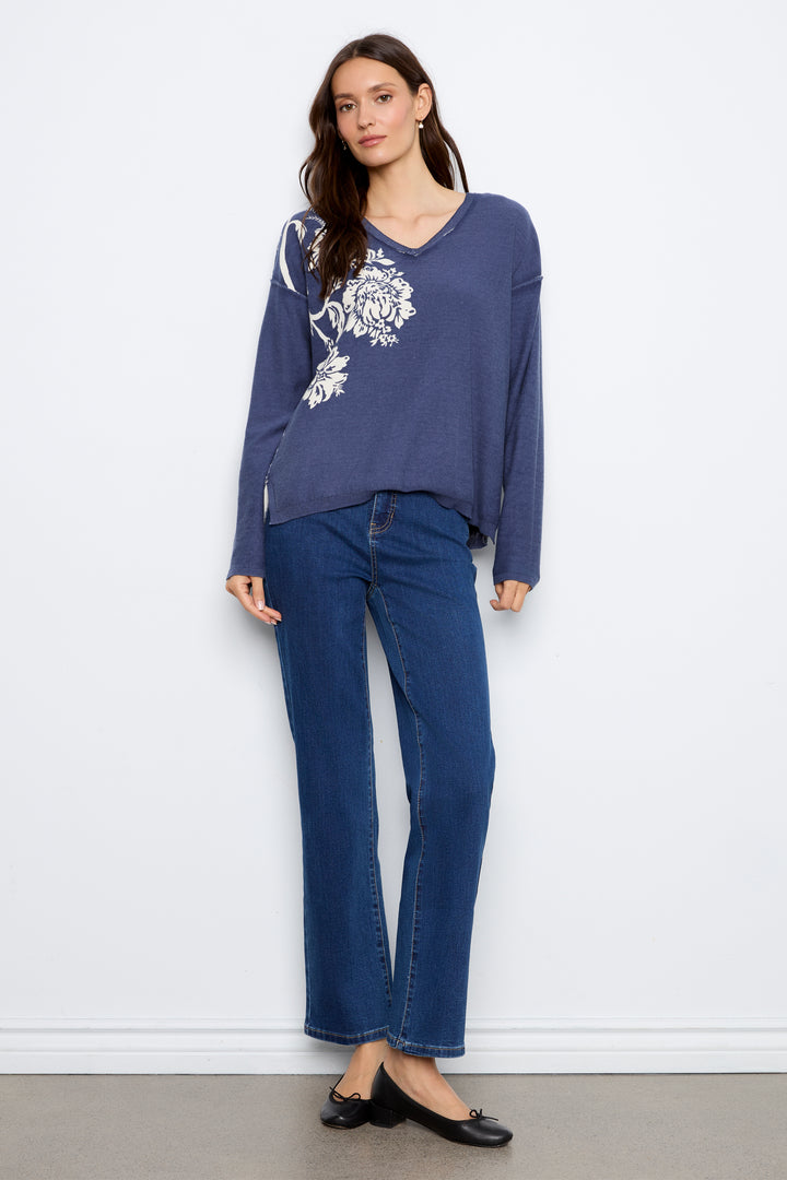 A woman modeling a slate blue long-sleeve knit top with white floral embroidery on the chest, paired with dark blue jeans and black flats, standing against a white wall.