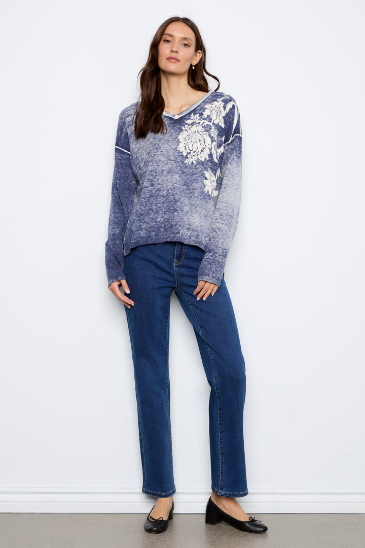 A woman stands against a white wall wearing a blue and white floral-patterned knit sweater, dark blue jeans, and black flats.