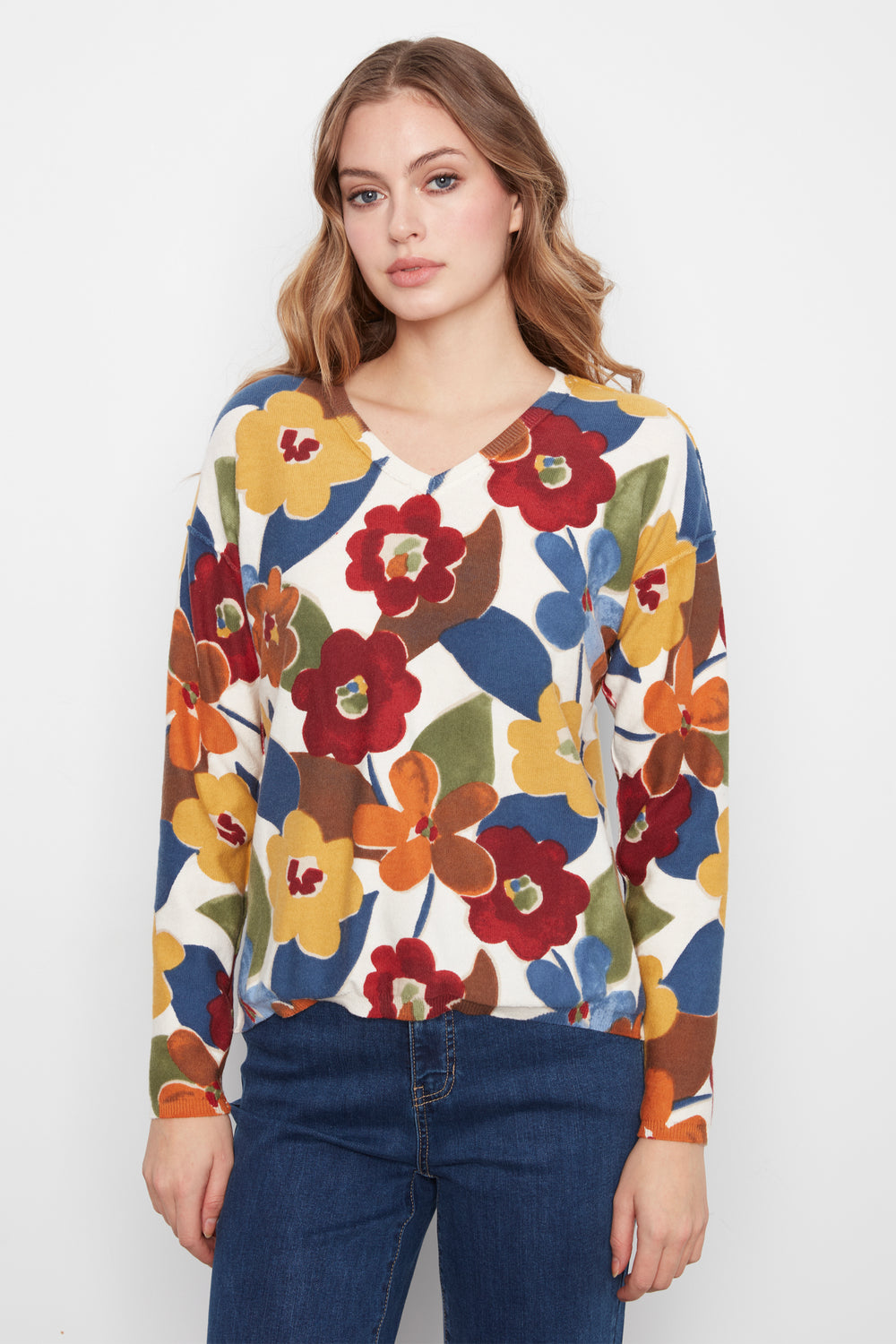 A woman wearing a long-sleeve V-neck top with a colorful floral pattern in red, yellow, orange, blue, and green on a white base, paired with dark blue jeans, against a plain light gray background.