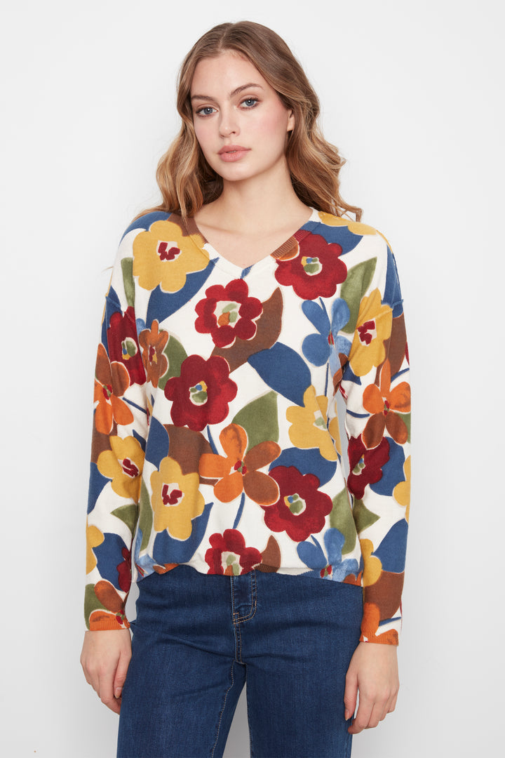 A woman wearing a long-sleeve V-neck top with a colorful floral pattern in red, yellow, orange, blue, and green on a white base, paired with dark blue jeans, against a plain light gray background.