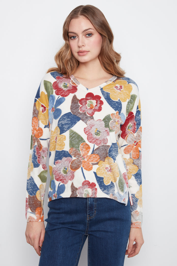 A woman wearing a long-sleeve V-neck top with a colorful floral print on a cream background, paired with dark blue jeans, against a plain light gray studio background.