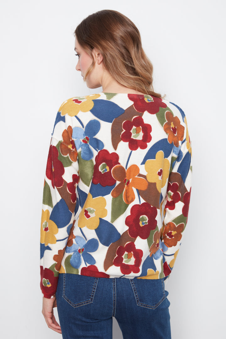 A woman viewed from the back wearing a cream-colored long-sleeve top with a vibrant, multicolored floral pattern in red, yellow, blue, and green, paired with dark blue jeans against a plain white background.