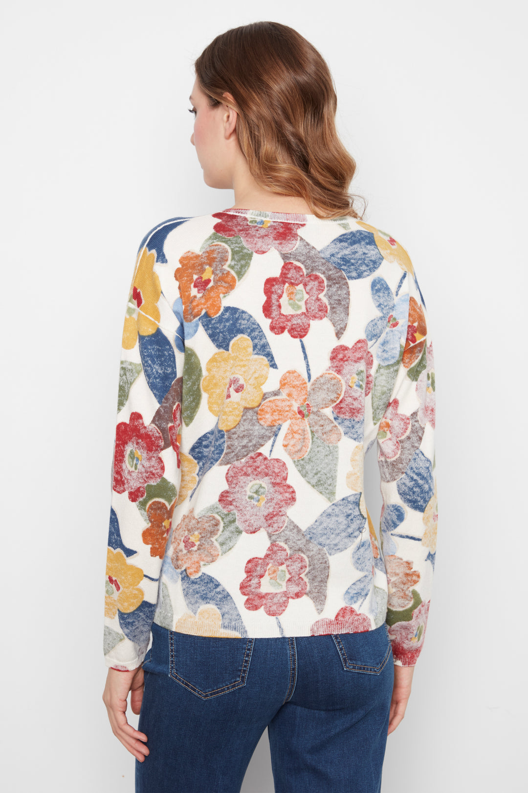 A woman viewed from the back wearing a cream-colored long-sleeve top with a vibrant, multicolored floral pattern in red, yellow, blue, and green, paired with dark blue jeans against a plain white background.
