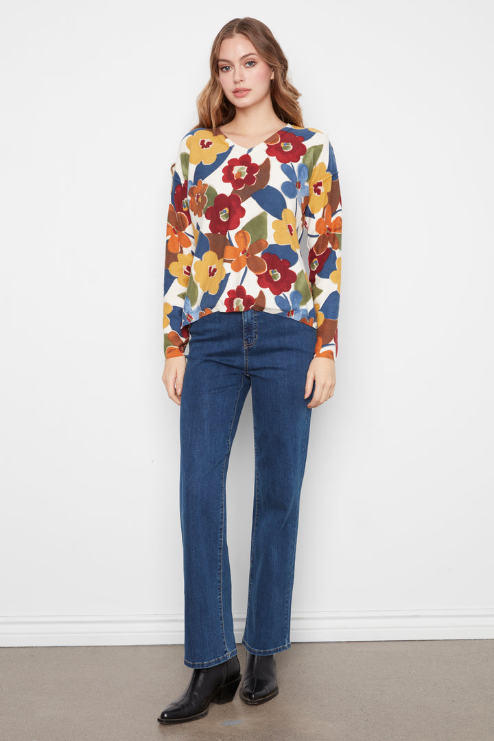 A woman modeling a long-sleeve V-neck top with a colorful floral pattern in red, yellow, blue, and green on a cream background, paired with dark blue jeans and black ankle boots, standing against a white wall.