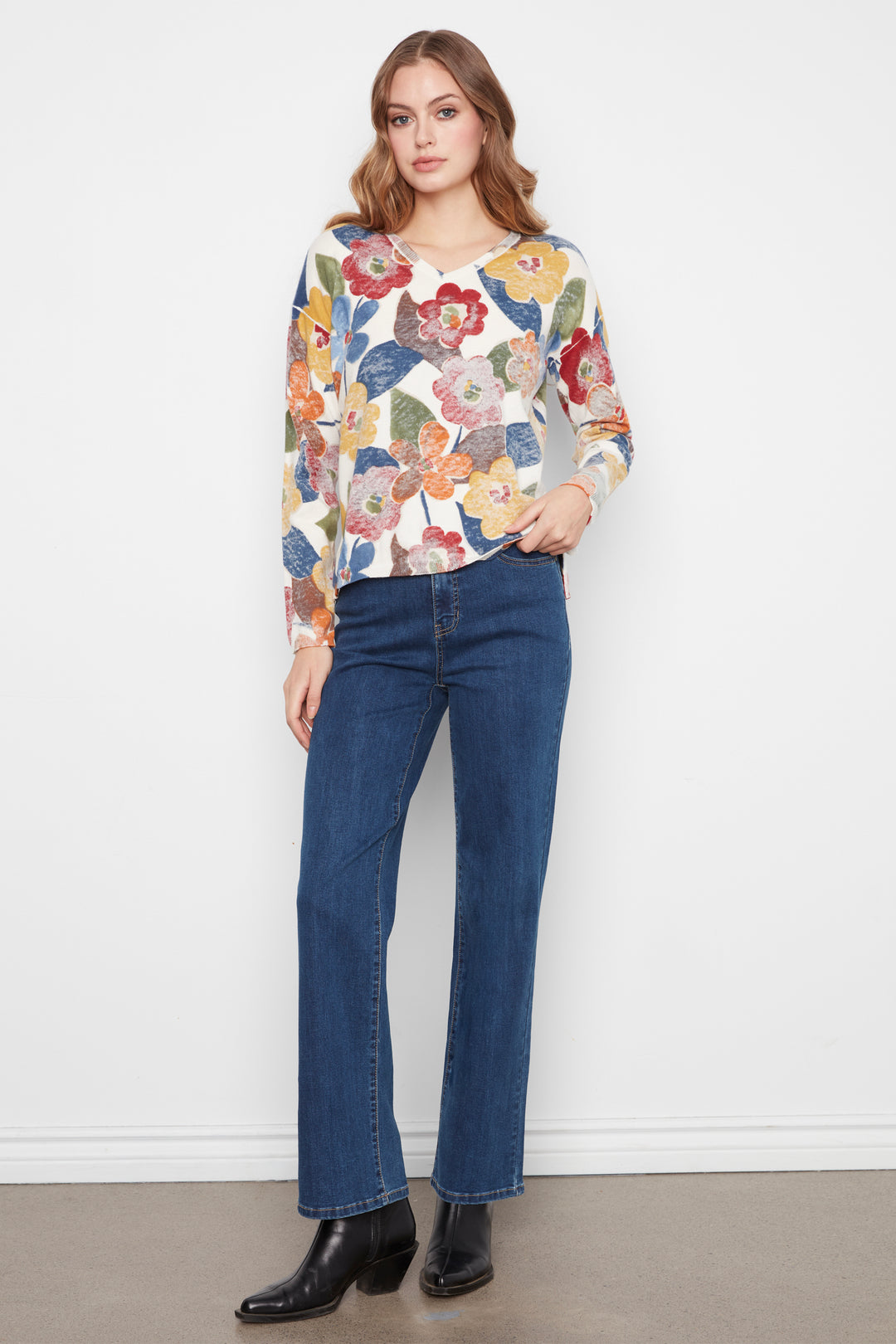 A woman modeling a long-sleeve V-neck top with a colorful floral pattern in red, yellow, blue, and green on a cream background, paired with dark blue jeans and black ankle boots, standing against a white wall.