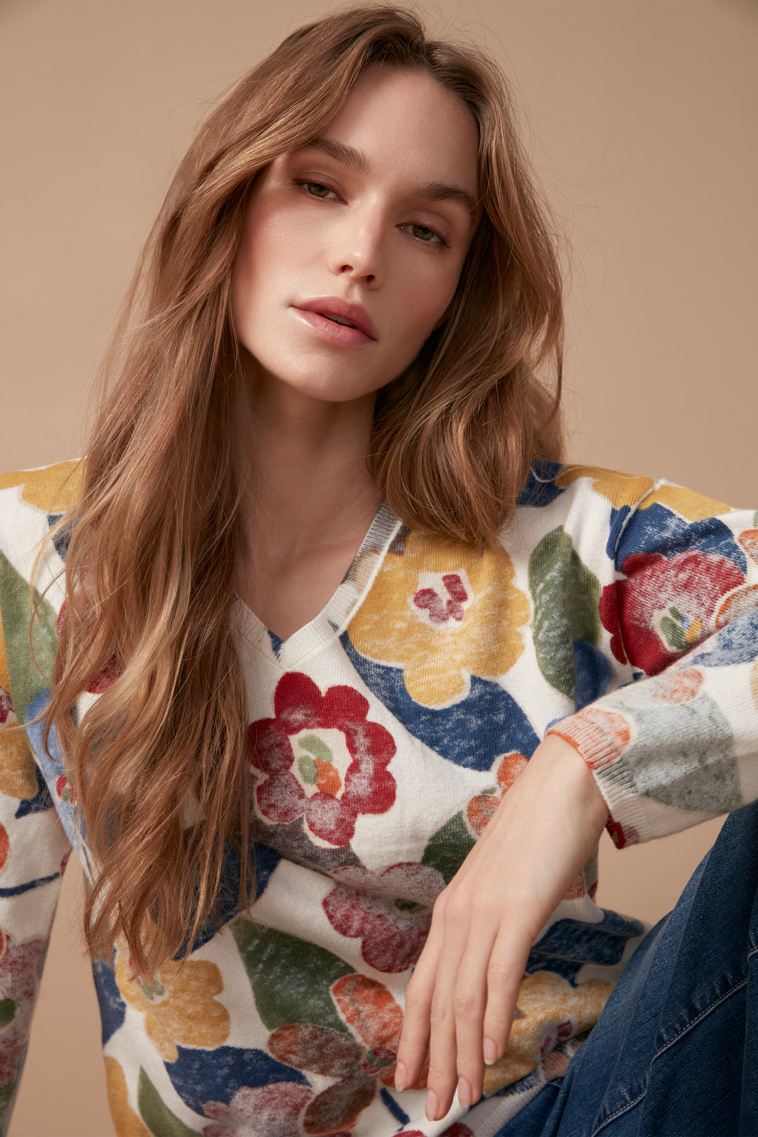 A woman wearing a long-sleeve V-neck top with a colorful floral pattern in red, yellow, orange, blue, and green on a white base, paired with dark blue jeans