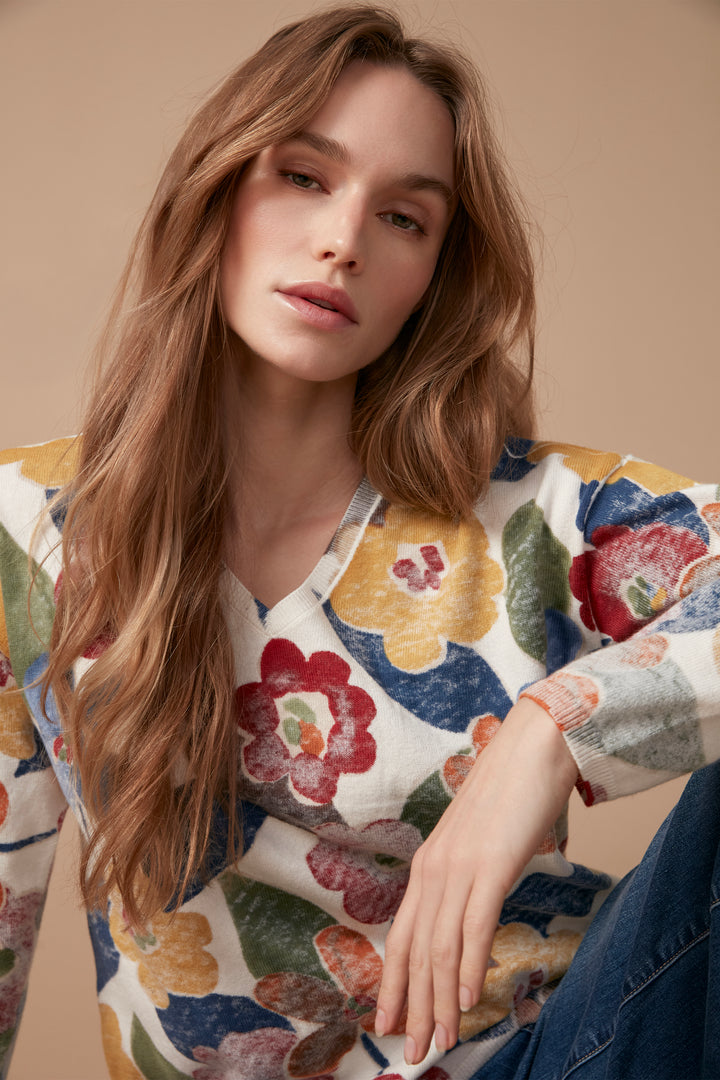 A woman wearing a long-sleeve V-neck top with a colorful floral pattern in red, yellow, orange, blue, and green on a white base, paired with dark blue jeans