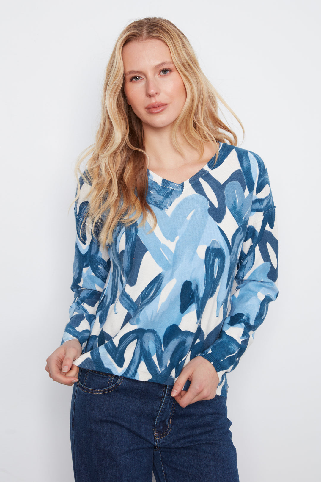 A woman with blonde hair wearing a blue and white abstract patterned long-sleeve top and dark blue jeans, standing against a light gray studio background.