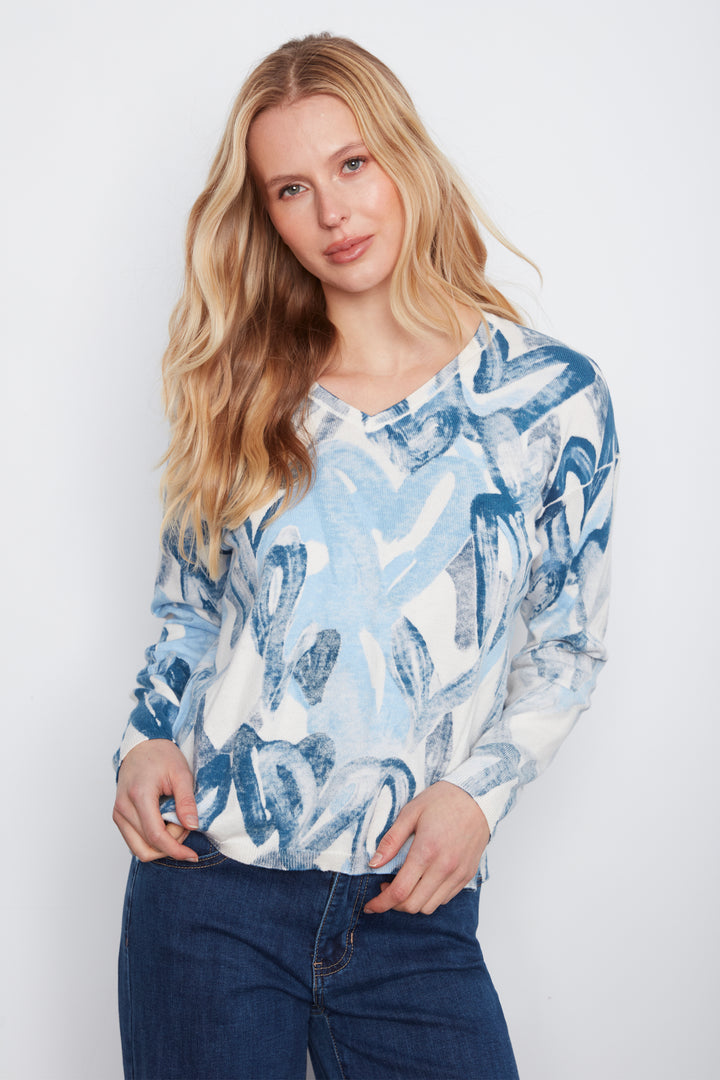 A woman with blonde hair wearing a light blue and white abstract patterned long-sleeve top and dark blue jeans, standing against a light gray studio background.