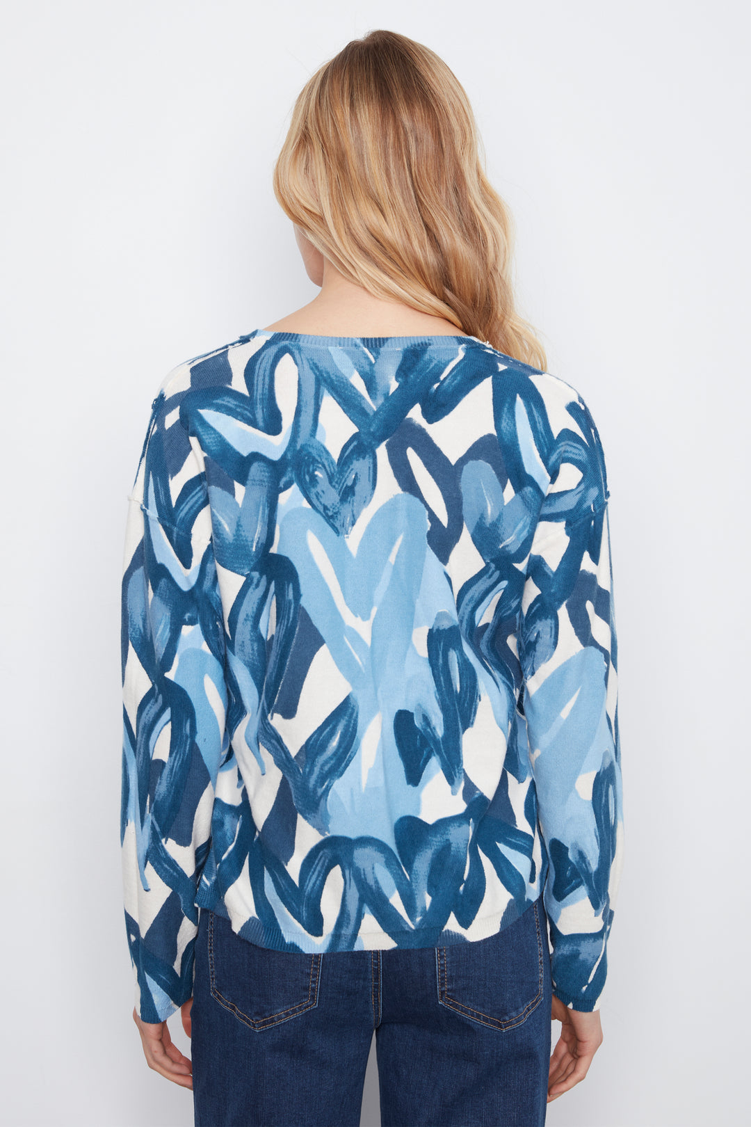 A woman viewed from behind wearing a long-sleeve top with an abstract blue and white brushstroke pattern, paired with dark blue jeans, against a plain white background.