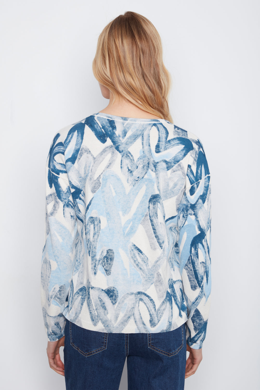 A woman viewed from behind wearing a long-sleeve top with an abstract blue and white brushstroke pattern, paired with dark blue jeans, against a plain white background.